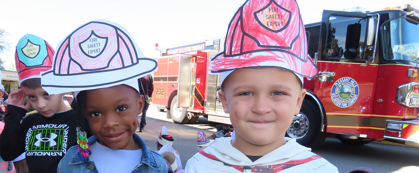 Two kindergarten students wear hats that say fire safety expert standing next to a fire truck